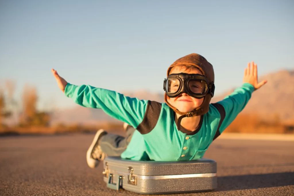 A child wearing aviator glasses and hat lays on top of a suitcase and pretends to be flying.