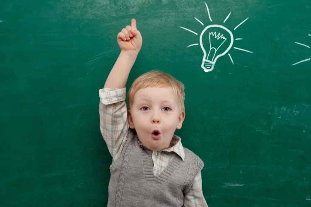 A child makes an excited face while pointing upwards. A lightbulb is written on the chalkboard behind the child.