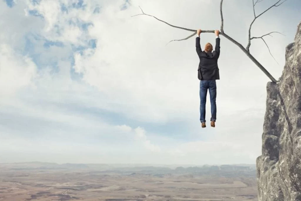 A person hanging precariously from a branch poking out the side of a mountain.