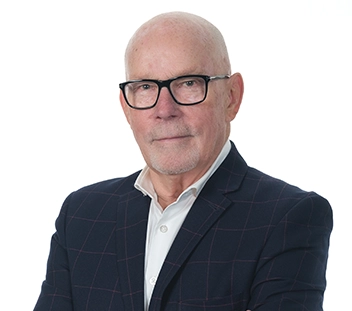 A person in a navy blue suit jacket and white button-up shirt wears black-rimmed glasses and looks towards the camera,