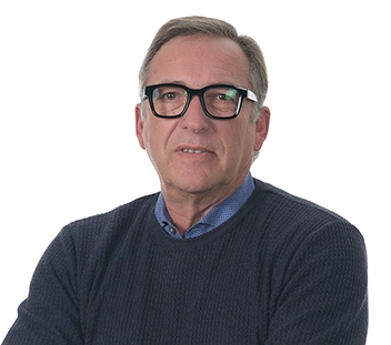 A person with short blong hair, wearing a navy blue sweater over a blue buttom-up and black-rimmed glasses looks toward the camera.