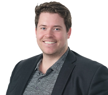 A person wearing a black suit jacket with a grey button up shirt smiles at the camera.