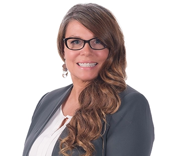A person with long reddish-brown hair, wearing black-rimmed glasses, a grey blazer, and white blouse smiles at the camera.